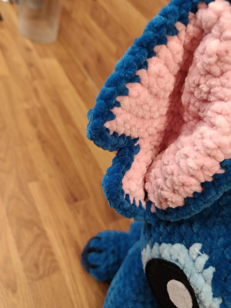 May include: A close-up of a blue and pink plush toy, featuring a large ear with a blue border and pink interior. The toy has a black and white eye and a blue body. The plush is made of a soft, textured material.