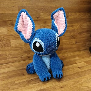 May include: A blue crochet plush toy of a cartoon character with large ears, black eyes, and a white belly. The ears have pink interiors. The toy is sitting on a wooden surface.