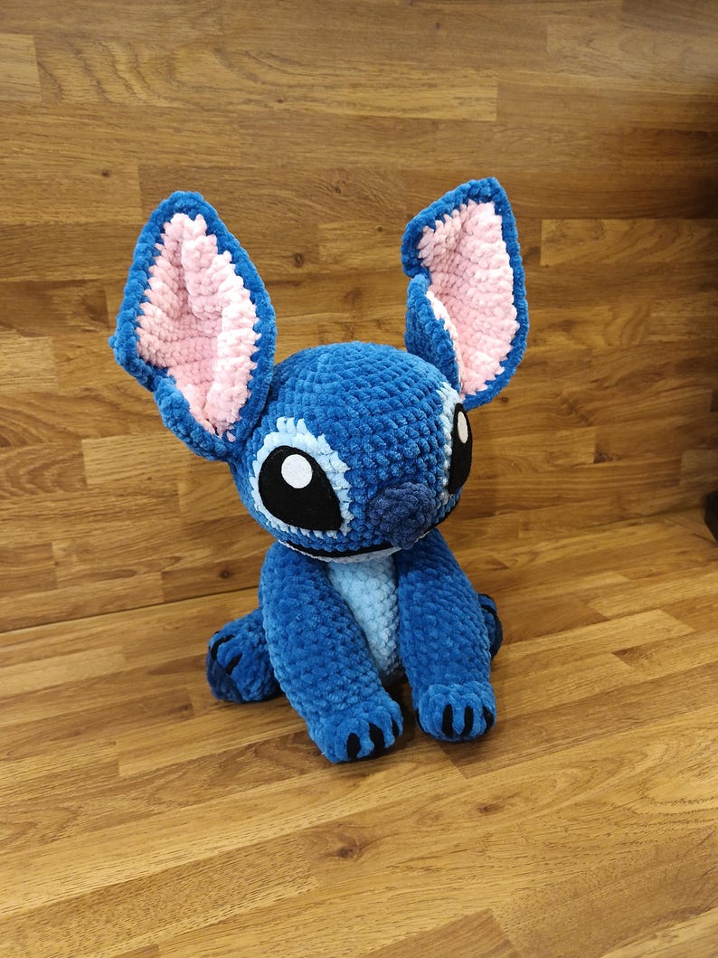 May include: A blue crochet plush toy of a cartoon character with large ears, black eyes, and a white belly. The ears have pink interiors. The toy is sitting on a wooden surface.