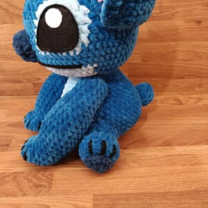 May include: A handmade blue crochet plush toy with large ears, a black eye, and a white and light blue eye detail. The plush has a small tail and black claws. The toy is sitting on a wooden surface.