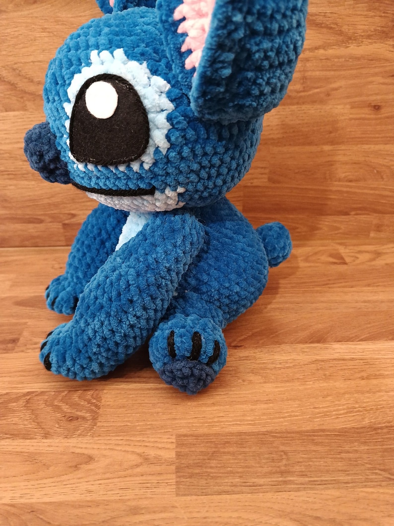 May include: A handmade blue crochet plush toy with large ears, a black eye, and a white and light blue eye detail. The plush has a small tail and black claws. The toy is sitting on a wooden surface.