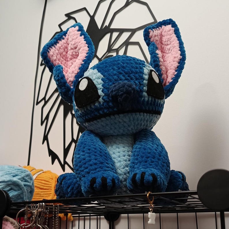 May include: A handmade blue crochet plush toy of a cartoon character with large ears, black eyes, and a light blue belly. The ears have pink inner linings. The plush is sitting on a black shelf.