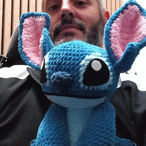 May include: A handmade blue crochet Stitch doll with pink inner ears and large black eyes. The plush toy is held in someone's hands. The doll is a popular character from a Disney animated film.