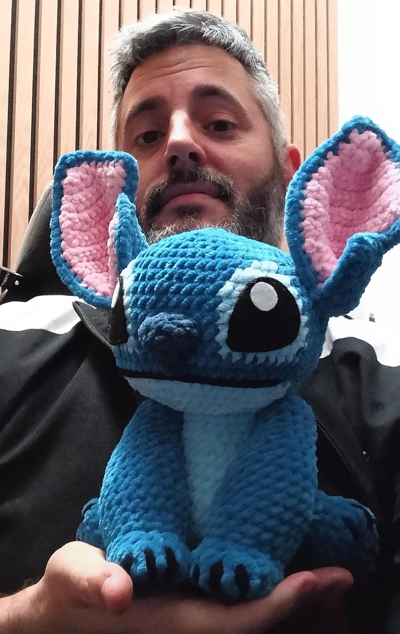 May include: A handmade blue crochet Stitch doll with pink inner ears and large black eyes. The plush toy is held in someone's hands. The doll is a popular character from a Disney animated film.