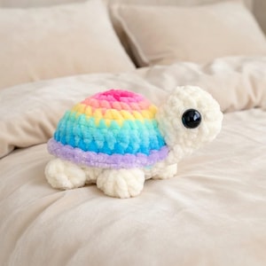 May include: A handmade crochet turtle plush toy. The turtle's shell is a vibrant rainbow of colors, including pink, yellow, blue, and purple. The body and head are white, with a large black eye. The plush toy is soft and cuddly.