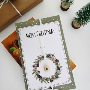 Pode incluir: Um cartão de Natal com a frase "MERRY CHRISTMAS" e um desenho de grinalda. O cartão está sobre um fundo verde com bolinhas brancas, com uma fita branca e uma pequena vela. Um presente embrulhado e árvores de Natal em miniatura também estão presentes.