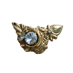 Puede incluir: Un broche de tono dorado con un diseño ornamentado que presenta un gran strass transparente en el centro y dos strass más pequeños en el lateral.