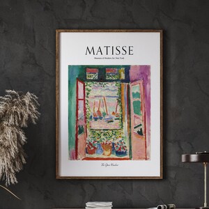 Op de afbeelding: Een ingelijste print van een schilderij van Henri Matisse getiteld "Het open raam". Het schilderij toont een kleurrijk uitzicht vanuit een raam met boten in de verte. De print is in een houten lijst en heeft de naam van de kunstenaar en het museum waar het schilderij zich bevindt afgedrukt onder de afbeelding.