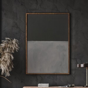 Pode incluir: Uma pintura abstrata enquadrada com uma metade superior cinza escura e uma metade inferior cinza clara. A pintura está pendurada em uma parede cinza escura em uma sala com uma mesa de madeira e uma lâmpada.