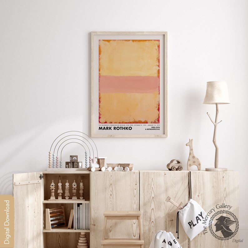 May include: A framed print of an abstract painting by Mark Rothko. The painting features a yellow background with a horizontal pink stripe. The print is hanging on a white wall in a room with wooden furniture and a lamp.