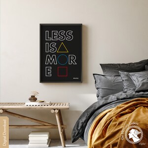 May include: A black and white typography print with the text "LESS IS MORE" in white with a yellow triangle, blue circle, and red square replacing the vowels. The print is framed in a black frame and hanging on a white wall.