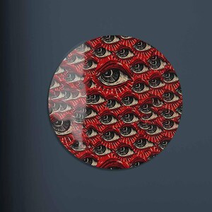 May include: A round wall art piece featuring a repeating pattern of stylized eyes on a red background. The eyes are black and white with radiating lines. The artwork is displayed on a dark blue wall above a teal sofa with orange and navy pillows.