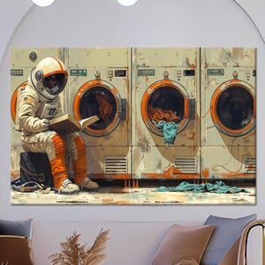 May include: A canvas print depicts an astronaut reading a book while sitting on a black container in front of three vintage washing machines. The astronaut wears a white and orange spacesuit. The machines have orange accents and contain colorful laundry.