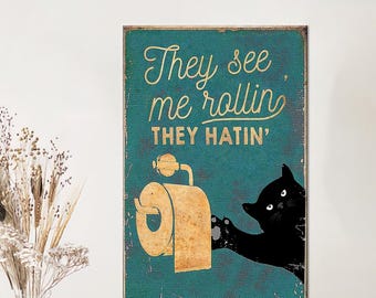 Könnte beinhalten: Türkis-goldenes Schild mit dem Text "They see me rollin', they hatin'". Eine schwarze Katze greift nach einer Toilettenpapierrolle. Das Schild hat einen verwitterten, Vintage-Look.