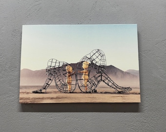 May include: Two wire-frame sculptures of people, one larger than the other, are standing in a desert landscape. The larger sculpture is holding the smaller sculpture in its arms.