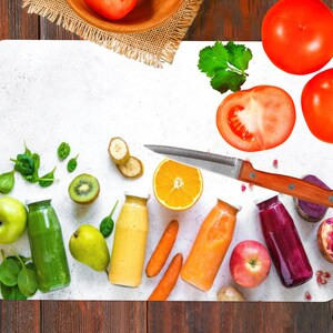 May include: A white cutting board with a variety of fruits and vegetables, including kiwi, bananas, oranges, apples, tomatoes, and spinach. There are also six glass bottles filled with different colored liquids, likely smoothies or juices. A silver knife with a wooden handle is placed on the board.
