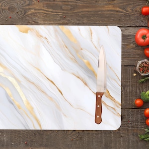 May include: Rectangular white cutting board with gold veining and a stainless steel knife with a wooden handle. The cutting board is on a wooden surface, surrounded by fresh vegetables and spices.