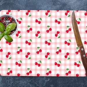 May include: A pink and white checkered cutting board with a repeating pattern of red cherries. A silver knife and a small bowl of spices are on the surface of the board.