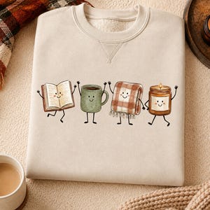 Könnte beinhalten: Ein beigefarbenes Sweatshirt mit Rundhalsausschnitt und einem Grafikdruck von vier anthropomorphen Figuren: ein offenes Buch, eine grüne Tasse, eine karierte Decke und eine brennende Kerze, jeweils mit lächelndem Gesicht sowie dünnen schwarzen Armen und Beinen.