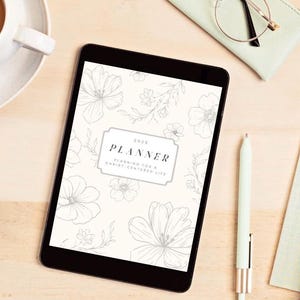 K&ouml;nnte beinhalten: Ein Tablet-Computer zeigt einen 2025-Planer mit floralen Illustrationen. Der Einband des Planers tr&auml;gt den Text "PLANNER" und "Planning for a Christ-centered life". Ein Stift, eine Brille und eine Tasse Kaffee sind ebenfalls auf der Holzoberfl&auml;che zu sehen.