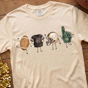 Könnte beinhalten: Ein cremefarbenes T-Shirt mit einer Grafik aus vier anthropomorphen Football-Gegenständen: einem braunen Football, einem schwarzen Trikot, einem beigen Helm und einem grünen Schaumstofffinger, alle mit lächelnden Gesichtern und Strichmännchen-Armen.