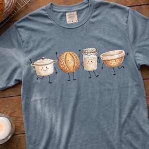 Könnte beinhalten: Ein schieferblaues Kurzarm-T-Shirt mit einem Grafikdruck von vier anthropomorphen Backutensilien: einem Schmortopf, einem runden Brotlaib, einem Glas mit Sauerteigstarter und einem Gärkörbchen, alle mit lächelnden Gesichtern und Strichbeinen.