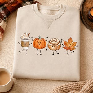 Könnte beinhalten: Ein beigefarbenes Sweatshirt mit Rundhalsausschnitt und einem Grafikdruck von vier anthropomorphen Figuren: einem Kaffeebecher mit Schlagsahne, einem orangefarbenen Kürbis, einer Zimtschnecke und einem herbstlichen Ahornblatt mit Gesichtern.