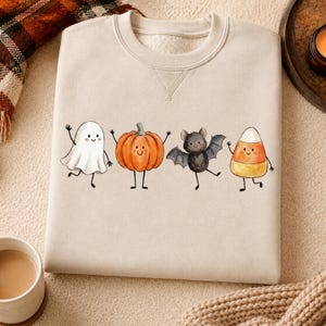 Könnte beinhalten: Ein beigefarbenes Sweatshirt mit Rundhalsausschnitt und einem Grafikdruck von vier Cartoon-Figuren in einer Reihe: ein weißes Gespenst, ein orangefarbener Kürbis, eine graue Fledermaus mit ausgebreiteten Flügeln und ein gelb-orange-weißes Zuckermais-Bonbon.