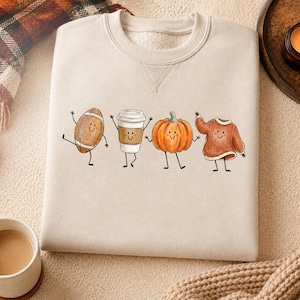 Op de afbeelding: Een beige sweater met ronde hals voorzien van een grafische print van vier antropomorfe figuren: een bruine football, een witte koffiebeker, een oranje pompoen en een roestkleurige kabeltrui, elk met dunne zwarte ledematen.
