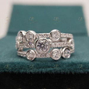 Puede incluir: Un anillo de plata con múltiples bandas, cada una adornada con piedras preciosas redondas y transparentes. El anillo presenta un diseño único con piedras más grandes engastadas en engastes de bisel, rodeadas de piedras de acento más pequeñas. El anillo se muestra sobre una superficie verde azulado.