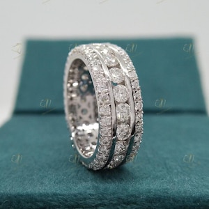 Puede incluir: Un anillo de diamantes de la eternidad con tres filas de diamantes de talla redonda. El anillo está engastado en un metal blanco, probablemente platino u oro blanco, y se muestra sobre una superficie de color verde azulado. El anillo brilla.