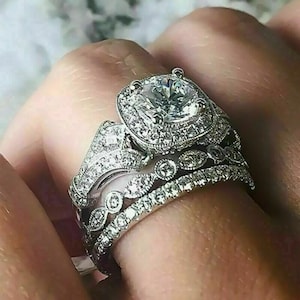 Peut inclure: Un ensemble de trois bagues en argent. La bague de fiançailles présente une grosse pierre précieuse ronde et transparente entourée de petites pierres. Les deux autres bagues ont des détails complexes et de petits pierres précieuses.