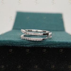 May include: A delicate, double-band ring with a row of small, clear diamonds on each band. The ring is made of white metal and is set against a green velvet background.