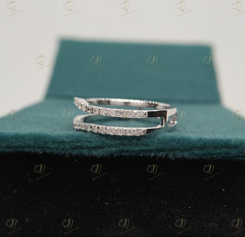 May include: A delicate, double-band ring with a row of small, clear diamonds on each band. The ring is made of white metal and is set against a green velvet background.
