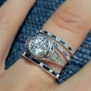 May include: A silver ring with a large, round, clear diamond center stone. The ring is surrounded by smaller diamonds and has a delicate band with blue sapphires. The ring is worn on a finger with three other bands, all with blue sapphires.