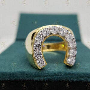May include: A gold ring with a horseshoe design set with clear round stones. The ring is displayed in a green velvet box.