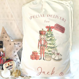 May include: A white Christmas gift bag with a drawstring closure, featuring a festive design. The bag displays a nutcracker, Christmas tree, and the words "Special Delivery for Jack." Red gift boxes and a rocking horse are also shown.