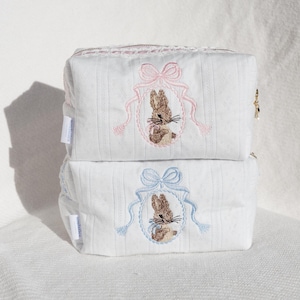Puede incluir: Dos bolsas de cosméticos de tela blanca con diseños de conejitos bordados. La bolsa superior tiene un lazo rosa y la bolsa inferior tiene un lazo azul.