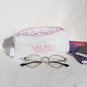 Puede incluir: Bolsa blanca con cremallera, forro floral rosa y el texto "Girl Math University" bordado en la parte delantera. La bolsa está sobre una superficie blanca con unas gafas de montura dorada y una pila de libros.