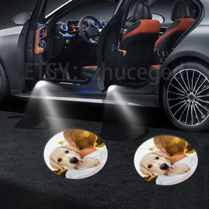May include: Two white LED car door lights project images of a golden retriever puppy being held by a person onto the ground. The puppy is looking at the camera with its tongue sticking out. The text "ETSY: hucege" is visible in the background.