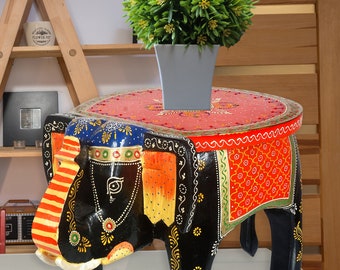 May include: A hand-painted wooden elephant stool with a black base and colorful accents. The stool has a round top and is decorated with intricate patterns. The elephant's trunk is raised and its eyes are painted with black and white details.