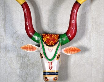 Pode incluir: Uma cabeça de touro de madeira branca com detalhes pintados em vermelho, amarelo, verde e azul. Os chifres são longos e curvos, e o rosto do touro é pintado com padrões intrincados.