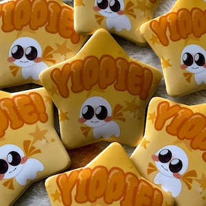 May include: Yellow star-shaped plush toys with the word "YIPPIE!" in orange lettering. Each star features a cartoon character with large eyes and a small bow. The background is a light colour.