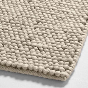 Puede incluir: Primer plano de una alfombra beige texturizada. La superficie de la alfombra está cubierta de pequeñas protuberancias redondeadas y elevadas, creando un efecto táctil y tridimensional. El color neutro y la textura densa sugieren un elemento de decoración hogareña acogedor y confortable.