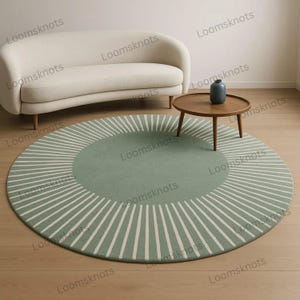 May include: A round, sage green rug with a white striped pattern radiating from the center. The rug is placed on a wooden floor, with a cream-colored sofa and a small wooden table with a vase in the background.