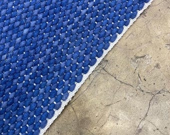 Puede incluir: Primer plano de una alfombra azul tejida con un borde blanco. La alfombra tiene un aspecto texturizado con un patrón de fibras entrelazadas. El color de la alfombra es un tono profundo de azul.