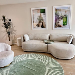 May include: A curved beige sofa with two matching throw pillows and a round green tray, positioned near a textured armchair, a tall potted plant, and a circular green rug with white radial lines, beneath two framed prints of Mediterranean scenes.