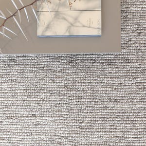 May include: A thick, textured, light gray wool rug with a looped pile surface. Resting on the rug is a smooth, light gray square table holding a Phaidon book titled "WORK" and a thin, thorny, light-colored branch casting a shadow.