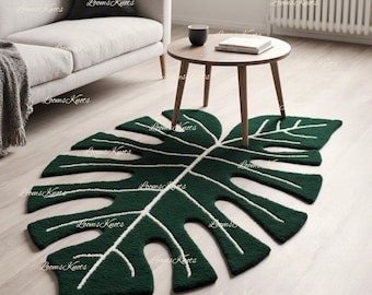 Puede incluir: Una alfombra verde oscuro con forma de hoja de monstera con detalles blancos. La alfombra está colocada sobre un suelo de madera clara. Una pequeña mesa redonda de madera y un sofá gris están en el fondo.
