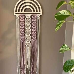 Könnte beinhalten: Ein Wandbehang aus Holz in Form eines Regenbogens mit einem Makrameemuster in Rosa- und Weißtönen.
